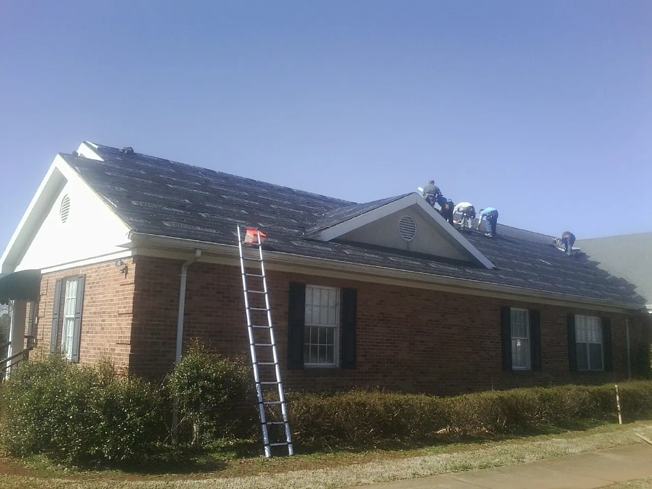 Crew tearing shingles off a brick ranch roof with a ladder set — Emergency Roof Repair work by our crew in Connelly Springs