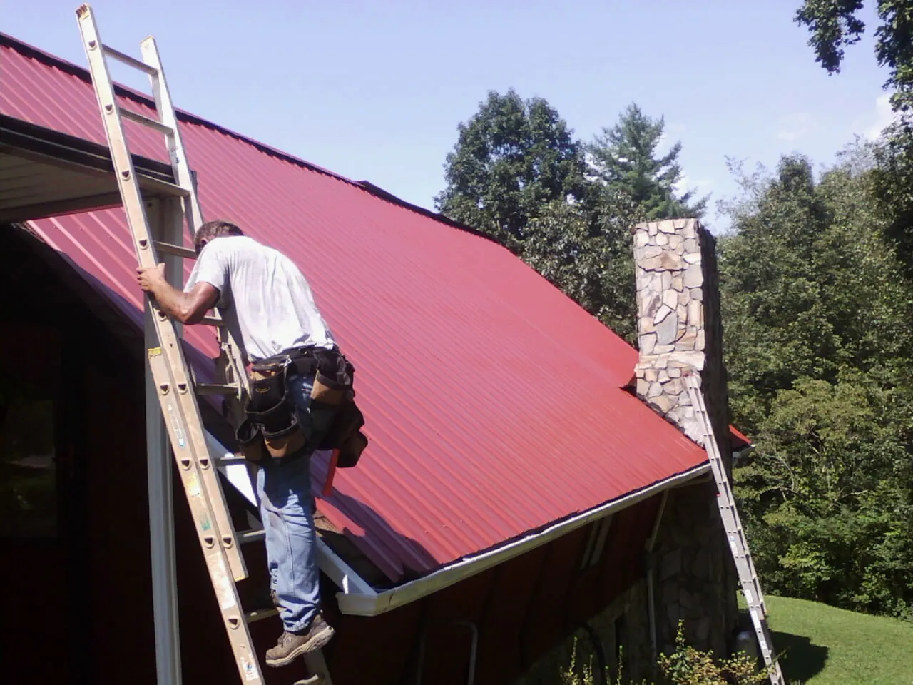 Roofer on a ladder beside a red metal roof and stone chimney — Soffit & Fascia Repair by Roofing Brothers in Connelly Springs